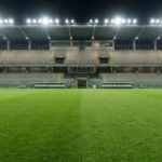 A green soccer pitch with mowing stripes faces an empty concrete grandstand illuminated by floodlights at night
